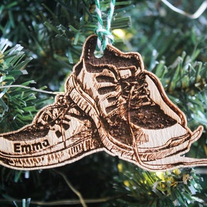 May include: A wooden Christmas ornament shaped like a pair of running shoes. The shoes are brown and have a white sole. The ornament is personalized with the name "Emma".