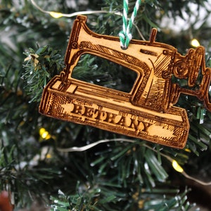 May include: A wooden Christmas bauble shaped like a vintage sewing machine with the name "Bethany" engraved on the base. The bauble is hanging on a green Christmas tree.