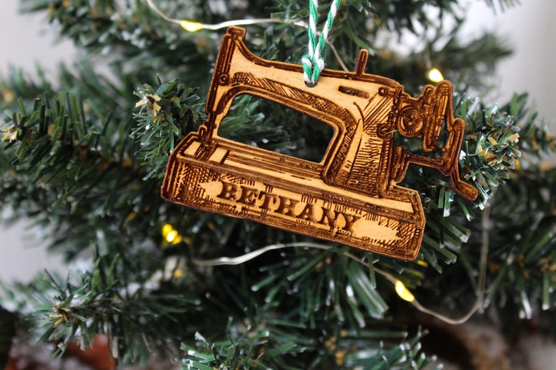 May include: A wooden Christmas bauble shaped like a vintage sewing machine with the name "Bethany" engraved on the base. The bauble is hanging on a green Christmas tree.