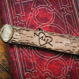 May include: A wooden bookmark with a heart design and the initials "A&J" carved into it. The bookmark is resting on a red book with a decorative pattern.