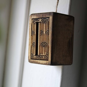 May include: A wooden block with the letter "L" carved into it. The letter is surrounded by a geometric pattern.