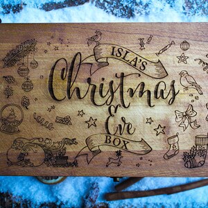 May include: A wooden Christmas Eve box with the inscription "Isla's Christmas Eve Box" and various Christmas-themed illustrations, including a snowman, a reindeer, a stocking, and a star.