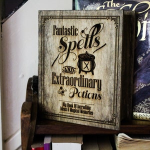 May include: A wooden book cover with a distressed woodgrain finish. The cover features a black and white design with the text "Fantastic Spells and Extraordinary Potions" and a cauldron with a cross. The text below the cauldron reads "Big Book of Incredible Ideas & Magical Memories".