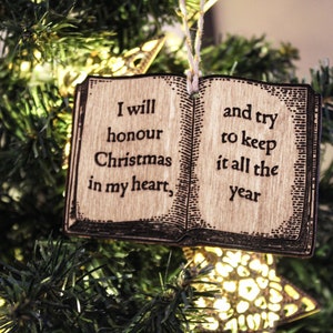 May include: Wooden Christmas ornament shaped like an open book, hanging on a Christmas tree. The ornament has the quote, "I will honour Christmas in my heart, and try to keep it all the year." The ornament is brown with black text.