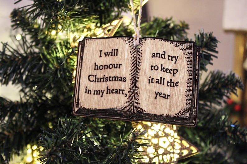 May include: Wooden Christmas ornament shaped like an open book, hanging on a Christmas tree. The ornament has the quote, "I will honour Christmas in my heart, and try to keep it all the year." The ornament is brown with black text.