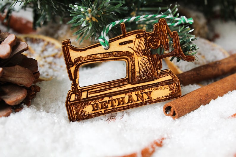 May include: A wooden Christmas bauble shaped like a vintage sewing machine with the name "Bethany" engraved on the bottom. The bauble is hanging from a green and white twine.