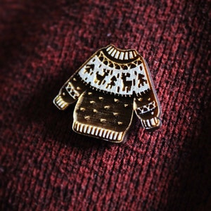 May include: A gold-colored metal pin shaped like a Christmas sweater with a white snowflake pattern and reindeer design.