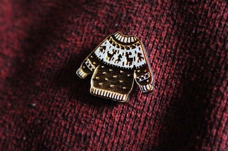 May include: A gold-colored metal pin shaped like a Christmas sweater with a white snowflake pattern and reindeer design.