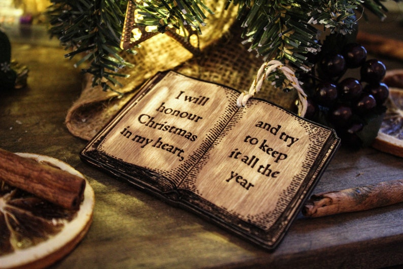 May include: Wooden Christmas ornament shaped like an open book, with the text "I will honour Christmas in my heart, and try to keep it all the year." The ornament is surrounded by festive decorations, including cinnamon sticks and greenery.