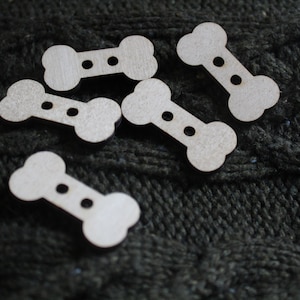 Könnte beinhalten: Fünf knopfartige Holzstücke in Knochenform mit jeweils zwei Löchern. Die Knöpfe sind hellbraun und auf einem dunkelgrauen Strickstoff angeordnet.