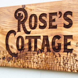 Könnte beinhalten: Ein Holzschild mit den Worten "Rose's Cottage", die in die Oberfläche gebrannt sind. Das Schild ist mit einem Rand aus kleinen Blumen und Schmetterlingen verziert.