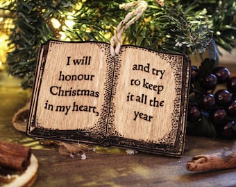 Peut inclure: Ornement de Noël en bois en forme de livre ouvert. L'ornement porte l'inscription gravée "I will honour Christmas in my heart, and try to keep it all the year". L'ornement est suspendu devant un sapin de Noël.