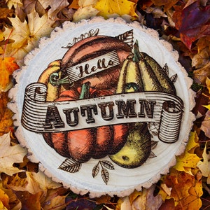 Può includere: Una fetta di legno con un'illustrazione disegnata a mano di zucche e pere. Il testo "Hello Autumn" è scritto su uno striscione.