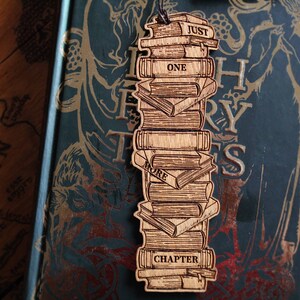 May include: Wooden bookmark shaped like a stack of books with the words "Just One More Chapter" on it. The bookmark is on top of a book with a green cover and gold lettering.