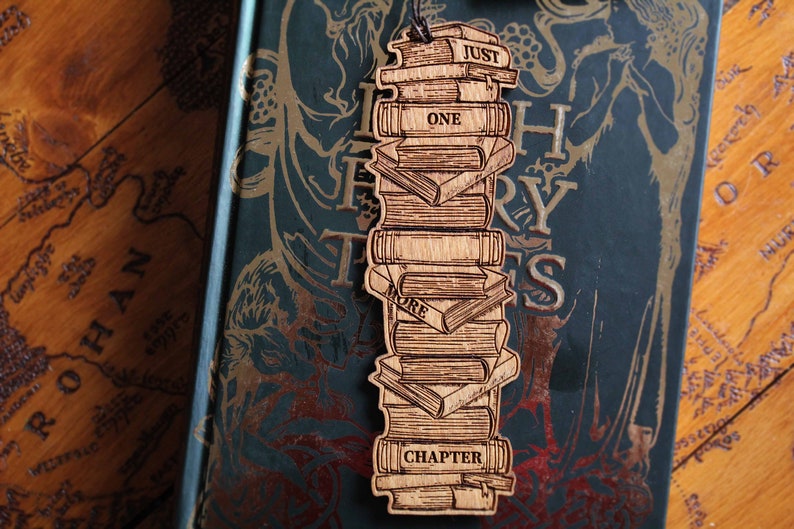 May include: Wooden bookmark shaped like a stack of books with the words "Just One More Chapter" on it. The bookmark is on top of a book with a green cover and gold lettering.