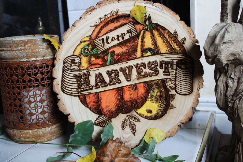 May include: A wooden round sign with a rustic wood grain texture. The sign features a hand-drawn illustration of pumpkins, gourds, and leaves. The text "Happy Harvest" is written in a decorative font.