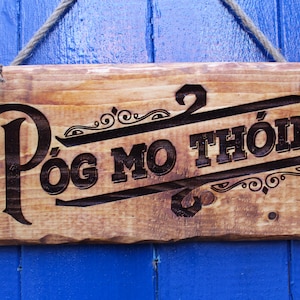 Può includere: Un cartello di legno con il testo "Póg Mo Thóin" in lettere nere. Il cartello è appeso a un muro blu.