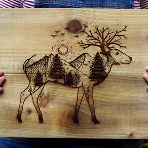 May include: A wood burned design of a deer with a mountain range and trees inside its body. The deer has antlers that are branches with leaves. The background is a light brown wood.