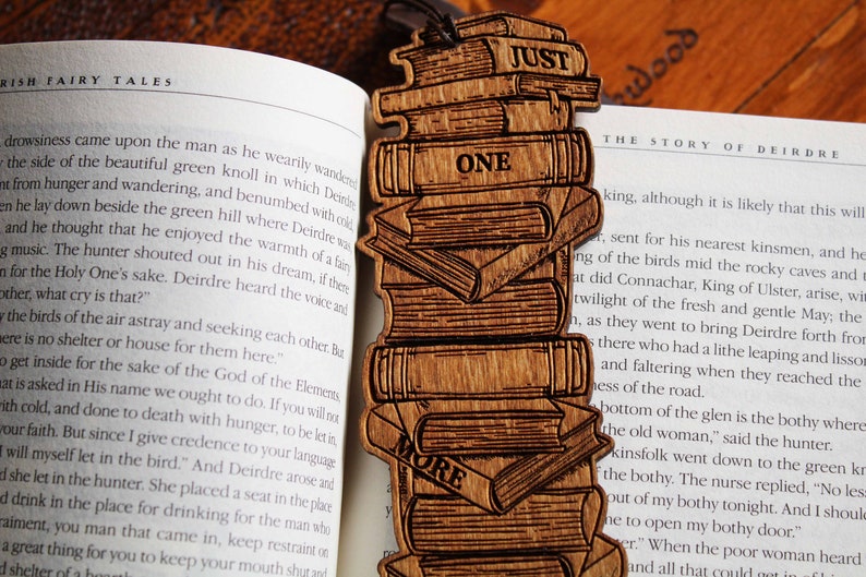 May include: A wooden bookmark with a stack of books engraved on it. The bookmark is brown and has the words "Just One More" engraved on it.