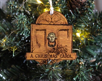 Op de afbeelding: Een houten kerstversiering in de vorm van een deur met een klopper in de vorm van een gezicht. De versiering is versierd met een takje hulst en de tekst "A Christmas Carol".