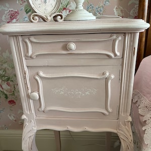 May include: A pink bedside table with a single drawer and a cabinet door. The table has ornate carvings and a distressed finish. It features a white clock with black Roman numerals and a white lamp with a white shade.