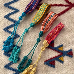 Könnte beinhalten: Vier bunte gewebte Armbänder mit Quasten. Die Armbänder sind aus verschiedenen farbigen Fäden gefertigt und haben ein geometrisches Muster. Die Quasten sind aus den gleichen farbigen Fäden wie die Armbänder.