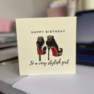 May include: A yellow card with the text "Happy Birthday" and a black and red illustration of a pair of high heels. The text "To a very stylish girl" is below the illustration.
