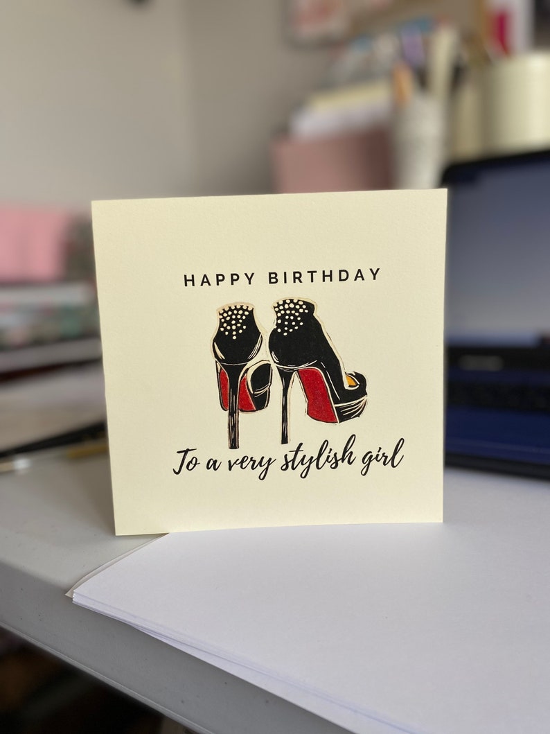 May include: A yellow card with the text "Happy Birthday" and a black and red illustration of a pair of high heels. The text "To a very stylish girl" is below the illustration.