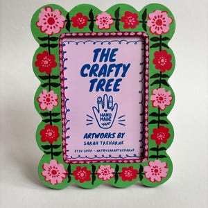 May include: A decorative picture frame with a scalloped green edge, adorned with pink and red floral designs. The frame's interior displays the text "THE CRAFTY TREE" and "ARTWORKS BY SARAH TREHARNE".