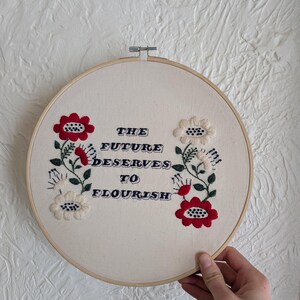 May include: Round embroidery hoop with cream-colored fabric featuring embroidered flowers and the text "THE FUTURE DESERVES TO FLOURISH" in navy blue. The flowers are red and white with green stems.