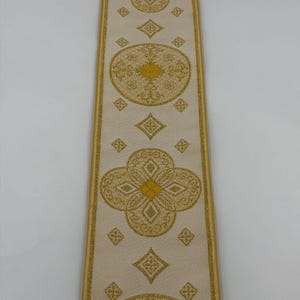 May include: Cream-colored ribbon with gold floral and geometric patterns. The ribbon features repeating quatrefoil and circular designs, with diamond shapes in between. The edges are trimmed with gold. The text "EMMA 253 No.1" is printed on the ribbon.