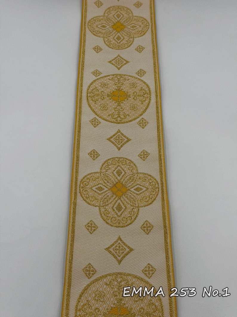 May include: Cream-colored ribbon with gold floral and geometric patterns. The ribbon features repeating quatrefoil and circular designs, with diamond shapes in between. The edges are trimmed with gold. The text "EMMA 253 No.1" is printed on the ribbon.