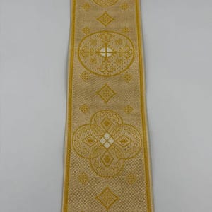 May include: A gold-colored ribbon with repeating ornate floral and geometric patterns. The ribbon has a textured appearance and features the text "EMMA 253 No.6" at the bottom. The design includes crosses and diamond shapes.