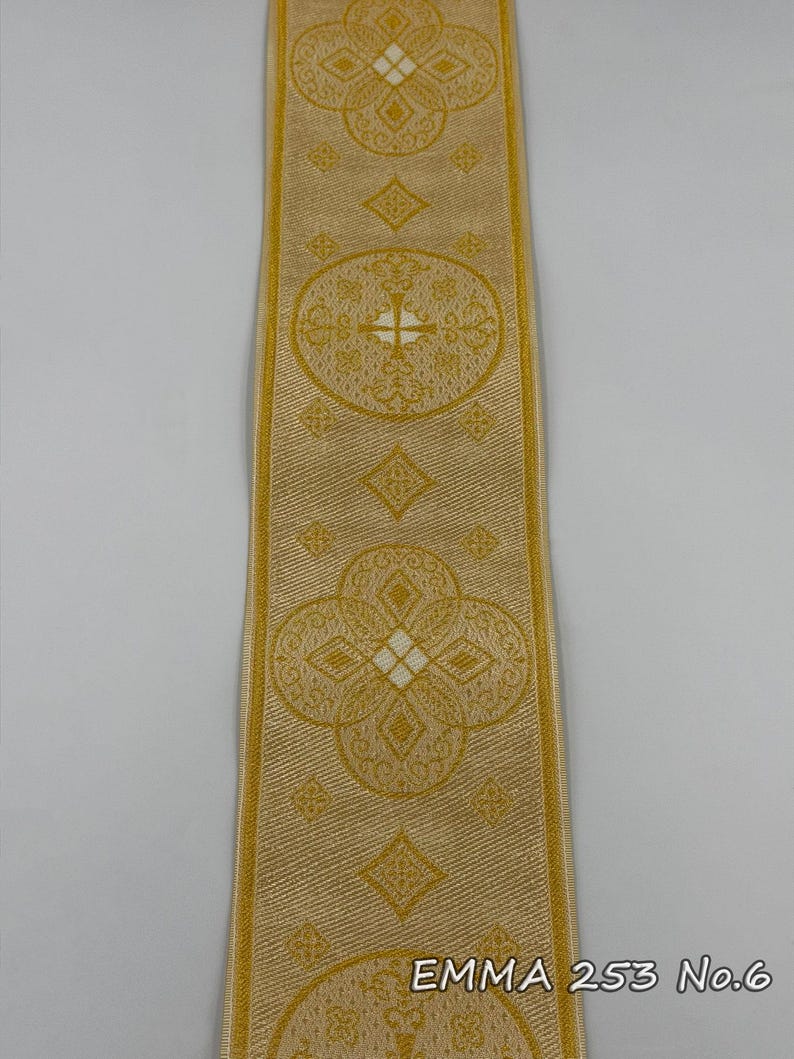 May include: A gold-colored ribbon with repeating ornate floral and geometric patterns. The ribbon has a textured appearance and features the text "EMMA 253 No.6" at the bottom. The design includes crosses and diamond shapes.