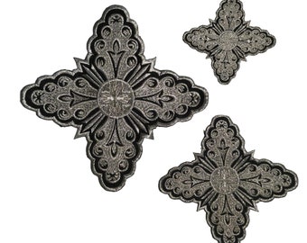 May include: Three silver and black embroidered cross patches of varying sizes. The crosses have a detailed design with intricate swirls and a center circle.