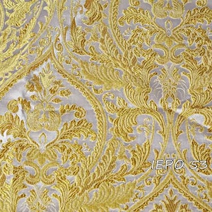 Pode incluir: Close-up de um tecido de damasco dourado e prateado com um padrão floral repetido. O tecido tem uma aparência texturizada com detalhes intrincados. O texto "IEPO 53 101" é visível no tecido.