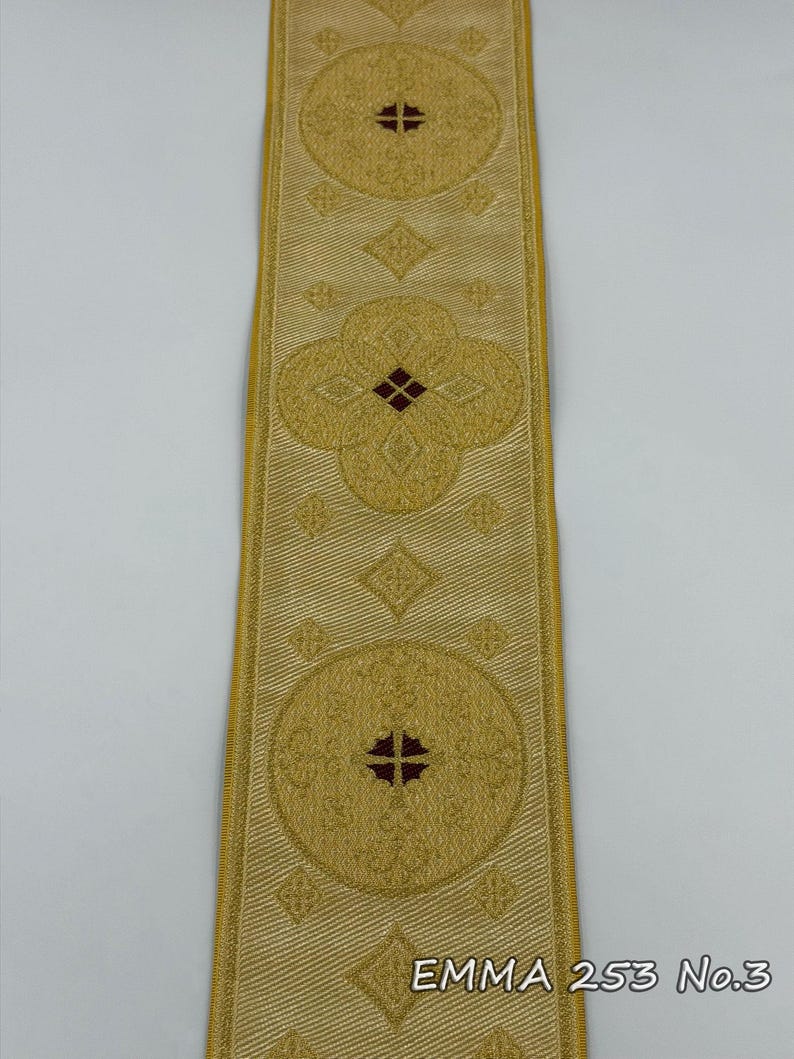 May include: Gold-colored decorative ribbon with repeating circular and floral patterns. The ribbon has a textured appearance and features a cross design in the center of the circles. The text "EMMA 253 No.3" is printed on the ribbon.