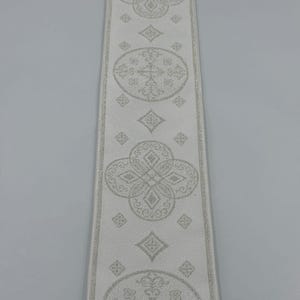 May include: A close-up of a white ribbon with a repeating pattern of floral and geometric designs in a lighter shade of white. The text "EMMA 253 No.7" is printed on the ribbon.