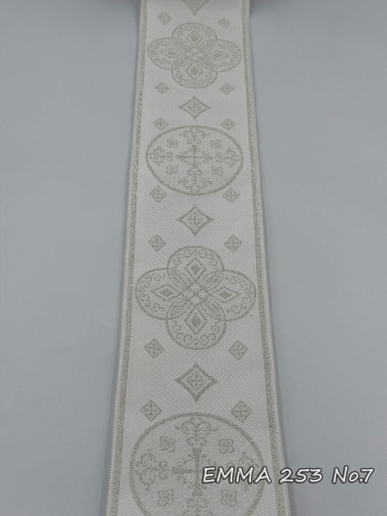 May include: A close-up of a white ribbon with a repeating pattern of floral and geometric designs in a lighter shade of white. The text "EMMA 253 No.7" is printed on the ribbon.