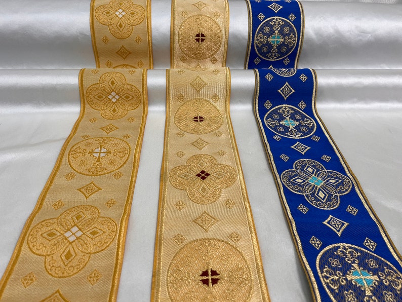 May include: Close-up of three decorative ribbons with intricate gold embroidery on a cream and blue background. The ribbons feature repeating patterns of crosses and floral designs, with gold trim.