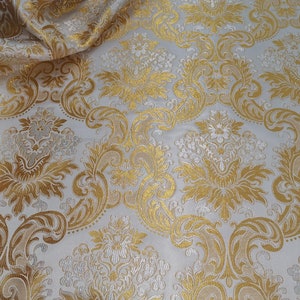 Könnte beinhalten: Weißer Stoff mit goldenem Damastmuster. Der Stoff hat eine erhabene Textur und ein luxuriöses Gefühl.
