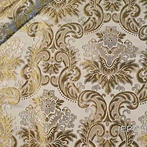 Pode incluir: Um close-up de um tecido branco com um padrão damasco dourado e prateado. O tecido tem um brilho sutil e uma sensação de luxo.