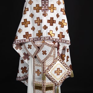 May include: White liturgical vestment with gold and maroon embroidery. The vestment features a pattern of crosses and intricate details.