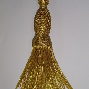 May include: A gold tassel with a woven top and a long fringe. The tassel is made of a shiny, metallic thread.