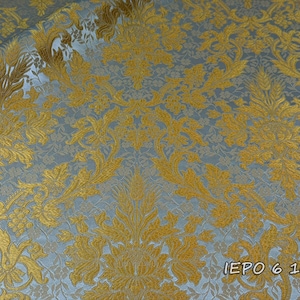 Pode incluir: Um tecido com um padrão de damasco em ouro metálico tecido sobre um fundo azul claro. O design intrincado consiste em motivos florais e de folhas simétricos. O texto IEPO 6 101 está impresso no canto inferior direito do tecido.