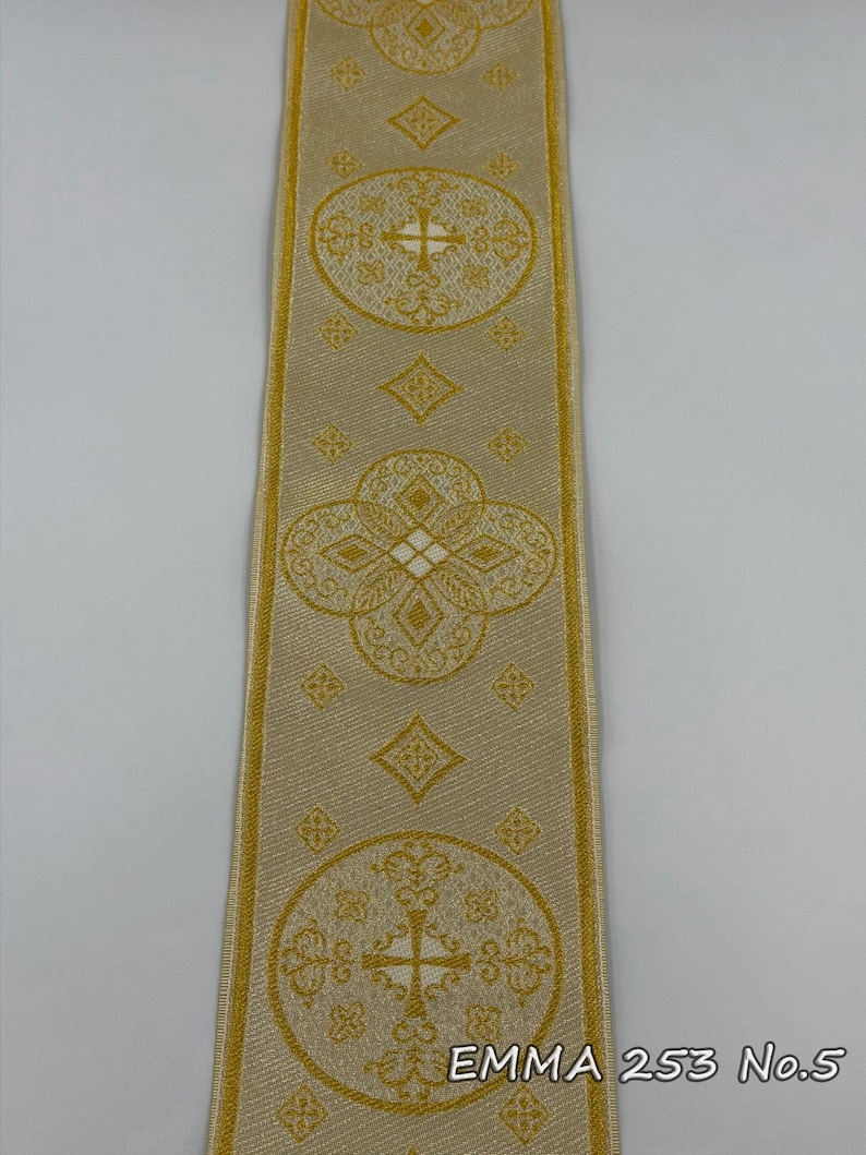 May include: A close-up of a decorative ribbon with a repeating pattern of gold crosses and floral designs on a beige background. The text "EMMA 253 No.5" is printed in the lower right corner.