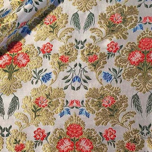 Pode incluir: Tecido marfim com um padrão repetitivo de desenhos florais e de folhas douradas, acentuados com flores vermelhas e azuis. O tecido tem uma aparência texturizada e um ligeiro brilho, adequado para estofados ou projetos decorativos.