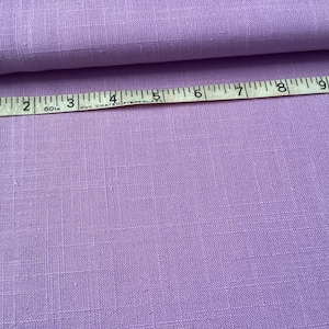 May include: A swatch of lavender fabric with a textured weave. A white and black measuring tape is laid across the fabric, showing measurements in inches. The fabric is folded over on itself.