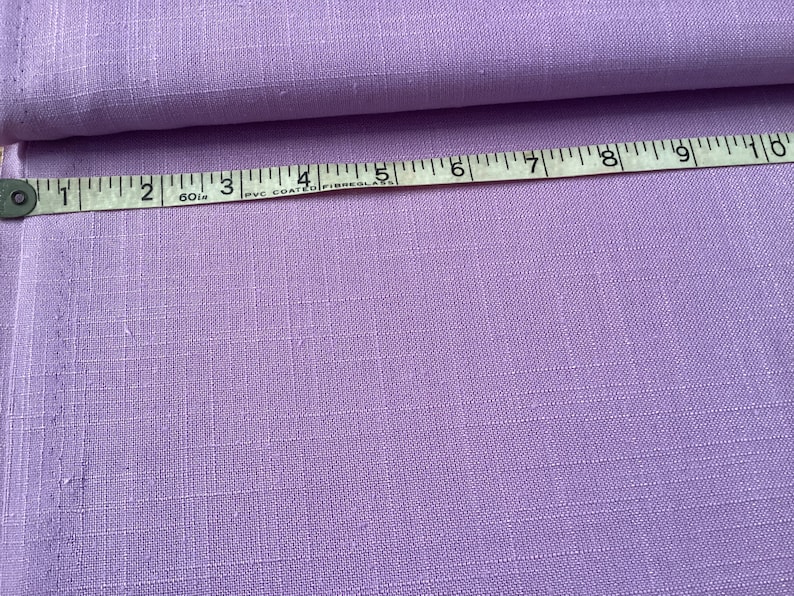 May include: A swatch of lavender fabric with a textured weave. A white and black measuring tape is laid across the fabric, showing measurements in inches. The fabric is folded over on itself.