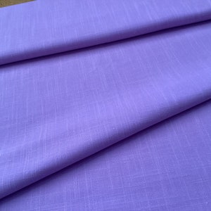 May include: A close-up of a folded piece of lavender fabric. The fabric has a subtle, textured weave, and the folds create shadows that add depth. The color is a soft, muted purple, and the fabric appears to be lightweight.